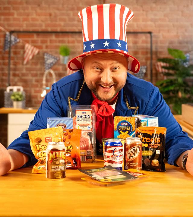 Sebastian Lege steht lachend hinter einer Küchentheke, auf der verschiedene Snacks aus den USA stehen. Er trägt einen Hut in den Farben der US-Flagge.
