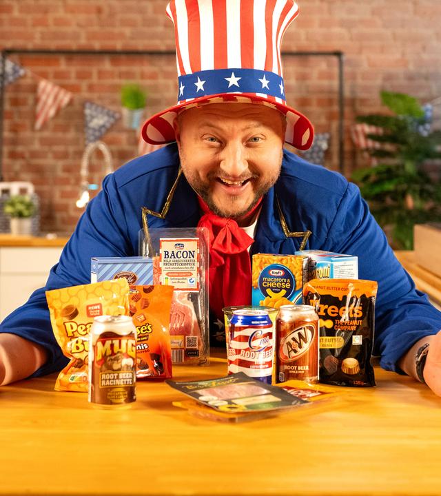Sebastian Lege steht lachend hinter einer Küchentheke, auf der verschiedene Snacks aus den USA stehen. Er trägt einen Hut in den Farben der US-Flagge.