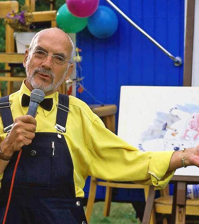 Peter Lustig in gelbem Hemd und Fliege präsentiert mit einem Mirkofon in der Hand vor dem Bauwagen ein gemaltes Bild.