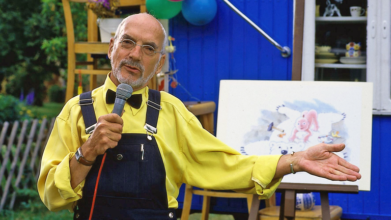 Peter Lustig in gelbem Hemd und Fliege präsentiert mit einem Mirkofon in der Hand vor dem Bauwagen ein gemaltes Bild.