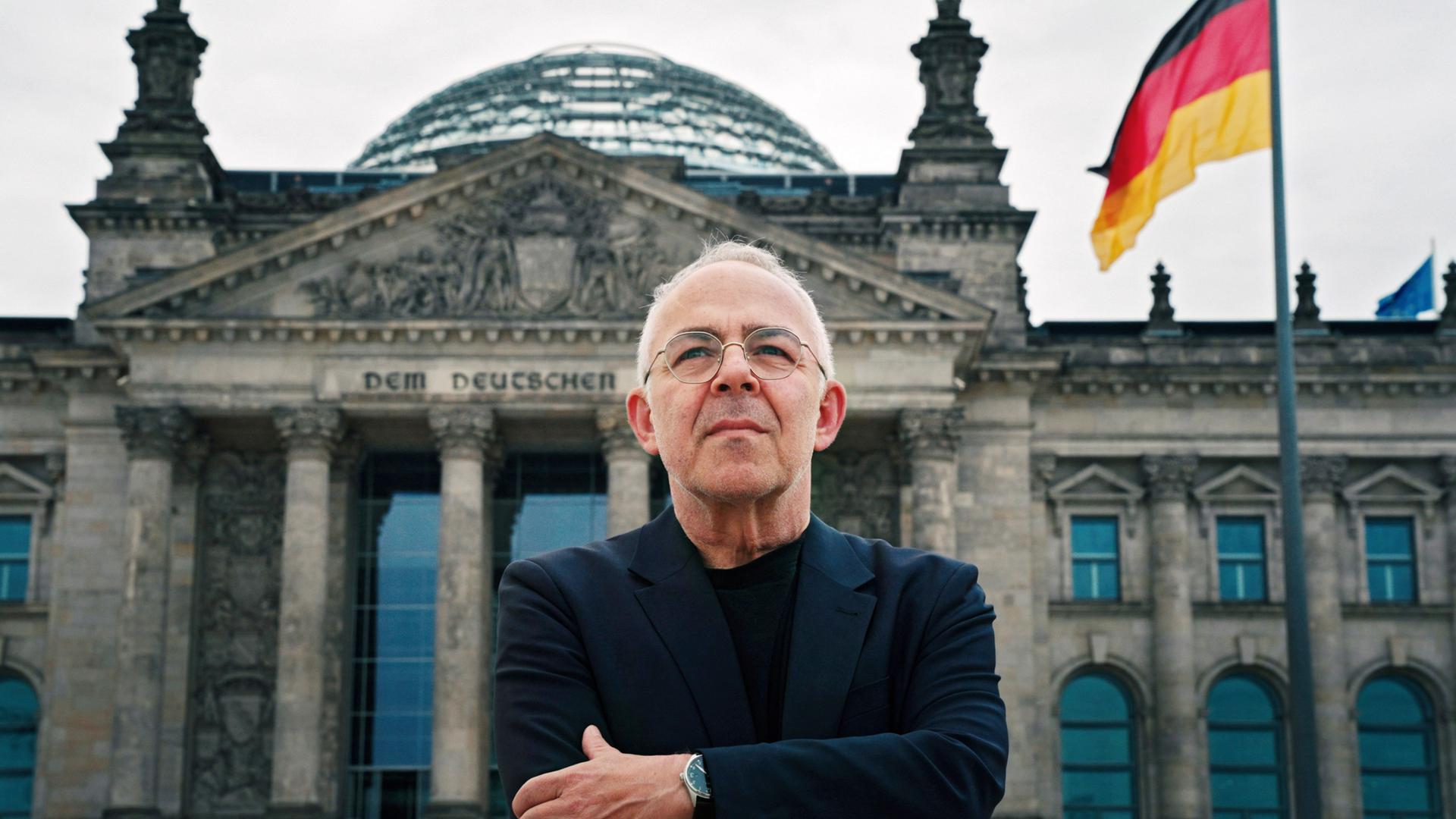 Ein älterer Mann mit Brille und dunkler Kleidung steht mit verschränkten Armen vor dem Reichstagsgebäude in Berlin, im Hintergrund weht eine deutsche Flagge.