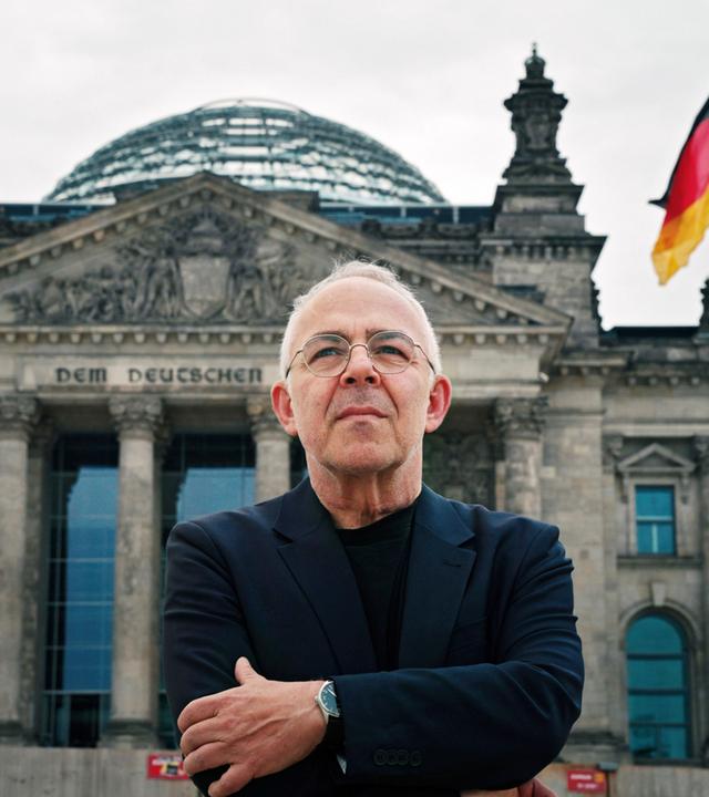 Ein älterer Mann mit Brille und dunkler Kleidung steht mit verschränkten Armen vor dem Reichstagsgebäude in Berlin, im Hintergrund weht eine deutsche Flagge.