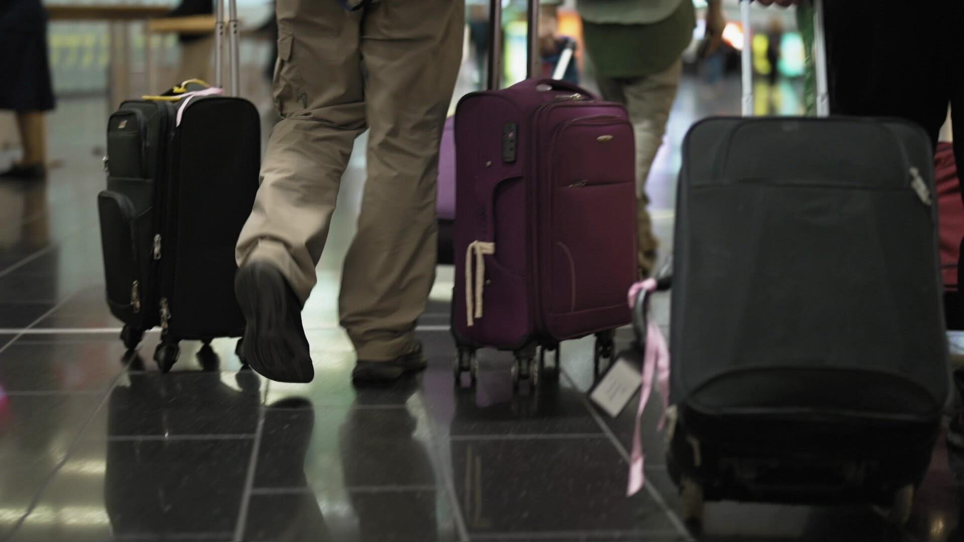 Das Bild zeigt eine Flughafen-Szene, in der mehrere Reisende mit Gepäck unterwegs sind. Im Vordergrund ist ein Mann zu sehen, der in beiger Kleidung läuft. Er zieht einen schwarzen Koffer mit sichtbaren Rollen hinter sich her. Neben ihm steht ein lila Koffer, ebenfalls mit Rollen, der mit einem pinkfarbenen Band versehen ist. Der Boden ist mit dunklen Fliesen ausgelegt, die einen glänzenden Effekt haben, und im Hintergrund sind verschwommene Figuren und Gepäck sichtbar, die ebenfalls zum Flughafenambiente gehören. Es scheint eine geschäftige Atmosphäre zu herrschen, die typisch für Reisende in einem Flughafen ist.