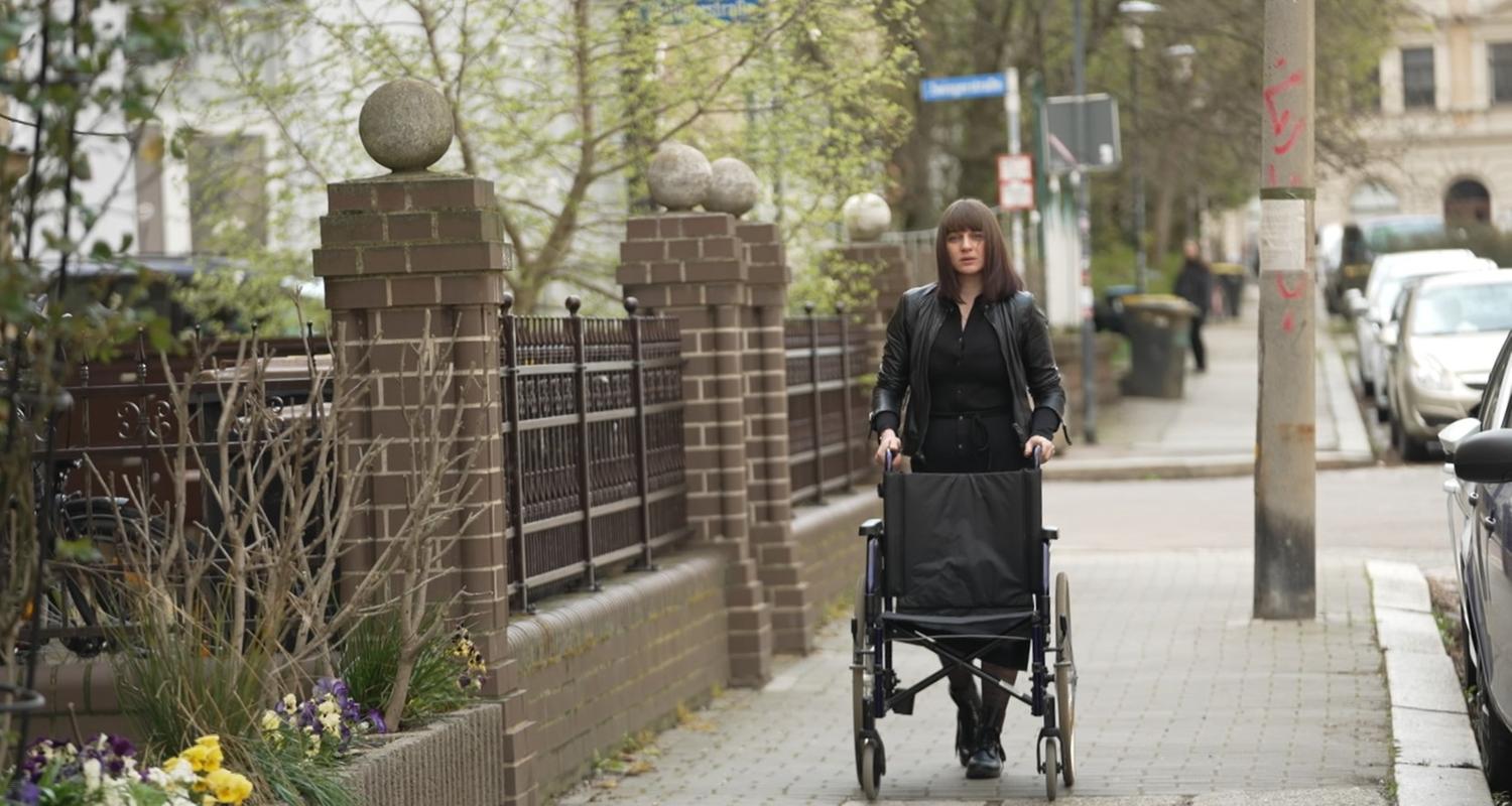 Betroffene von Folgen nach-Corona-Impfung, die ihren Rollstuhl auf einem Gehweg schiebt