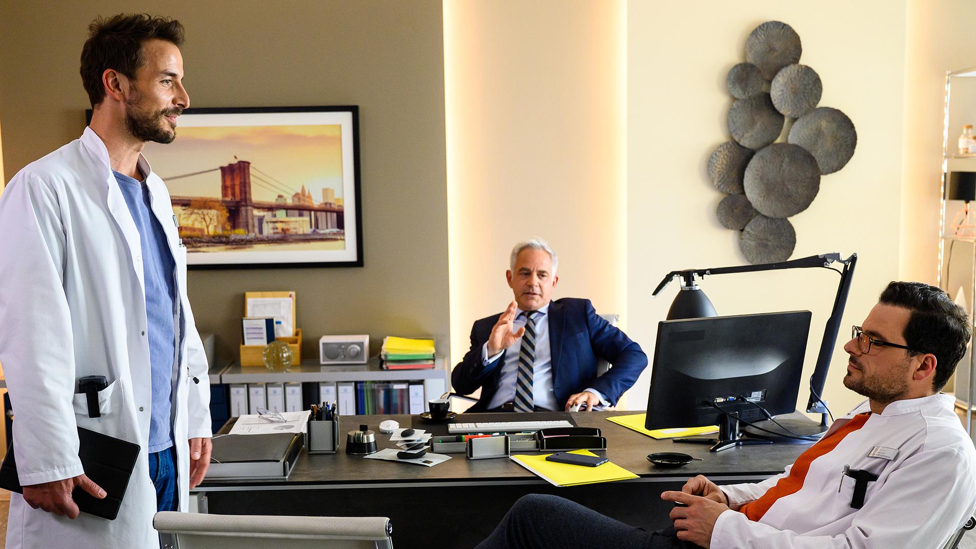 Dr. Frank Stern (Max Alberti) steht in Prof. Dr. Alexander von Arnstetts (Florian Fitz) Büro und spricht mit dem, auf einem Stuhl sitzenden, Dr. Tom Koopmann (Tobias Licht). Dr. von Arnstett beobachtet die Szene von seinem Schreibtischstuhl aus.
