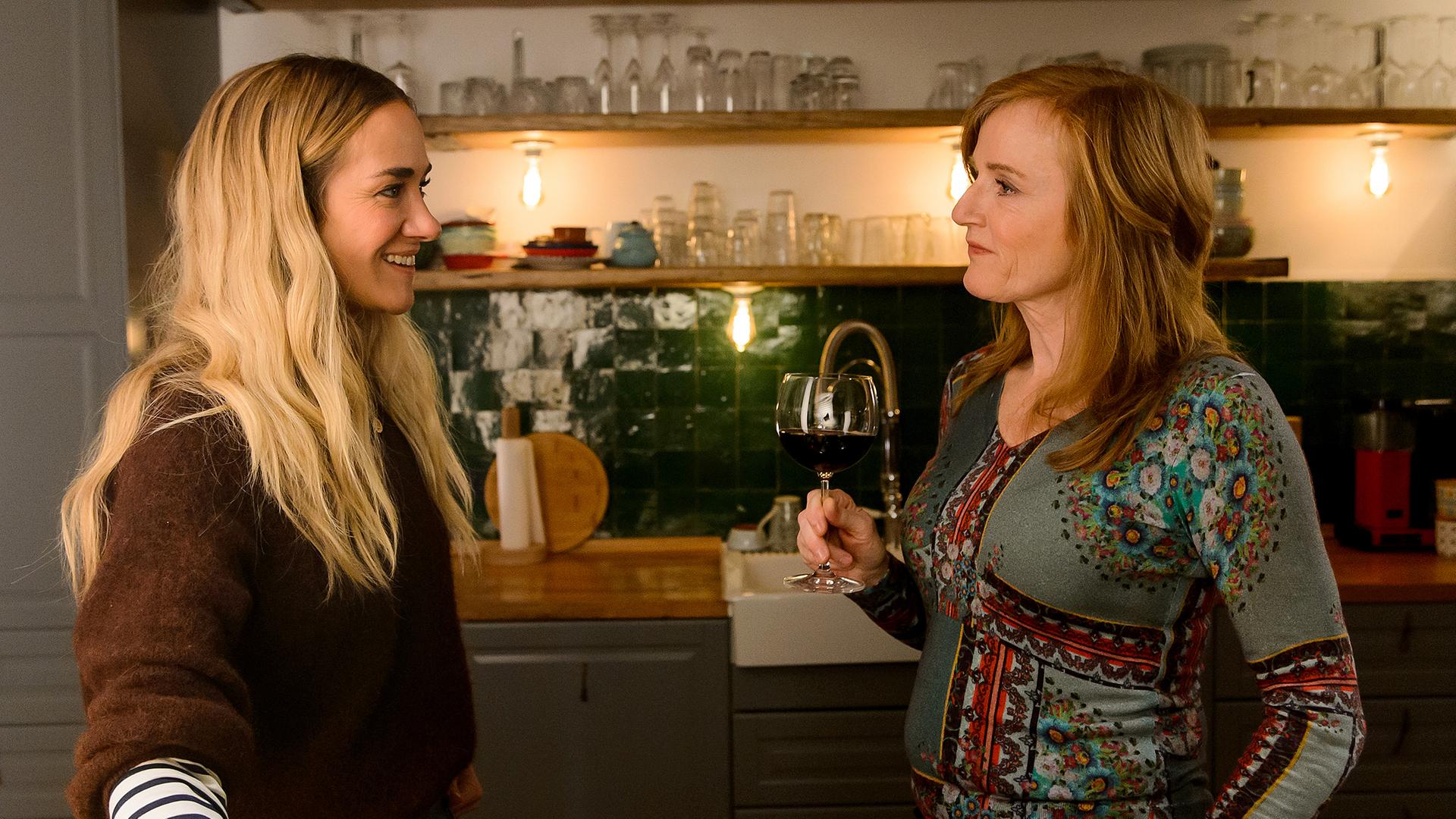 Betty (Annina Hellenthal) und ihre Mutter Gabriele (Nina Petri) stehen sich in Bettys Küche gegenüber. Gabriele hält ein Glas Wein und lächelt Betty an, die glücklich zurück lächelt.
