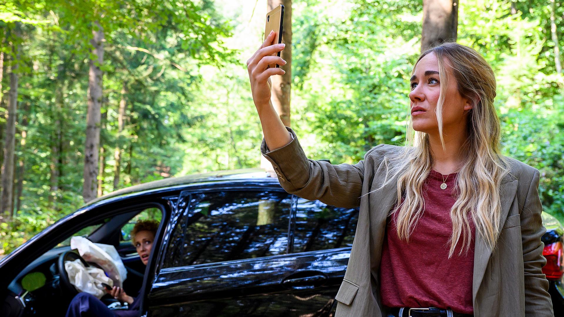 Dr. Helena von Arnstett (Claudia Hiersche) sitzt auf dem Fahrersitz im Auto und schaut Betty (Annina Hellenthal) dabei zu, wie sie ihr Handy in die Luft hält um im Wald nach einem besseren Empfang zu suchen.
