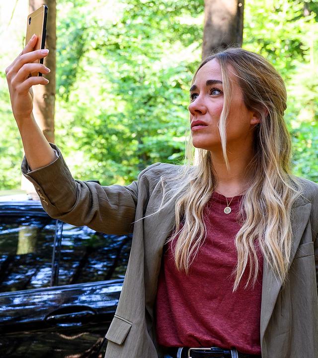 Dr. Helena von Arnstett (Claudia Hiersche) sitzt auf dem Fahrersitz im Auto und schaut Betty (Annina Hellenthal) dabei zu, wie sie ihr Handy in die Luft hält um im Wald nach einem besseren Empfang zu suchen.