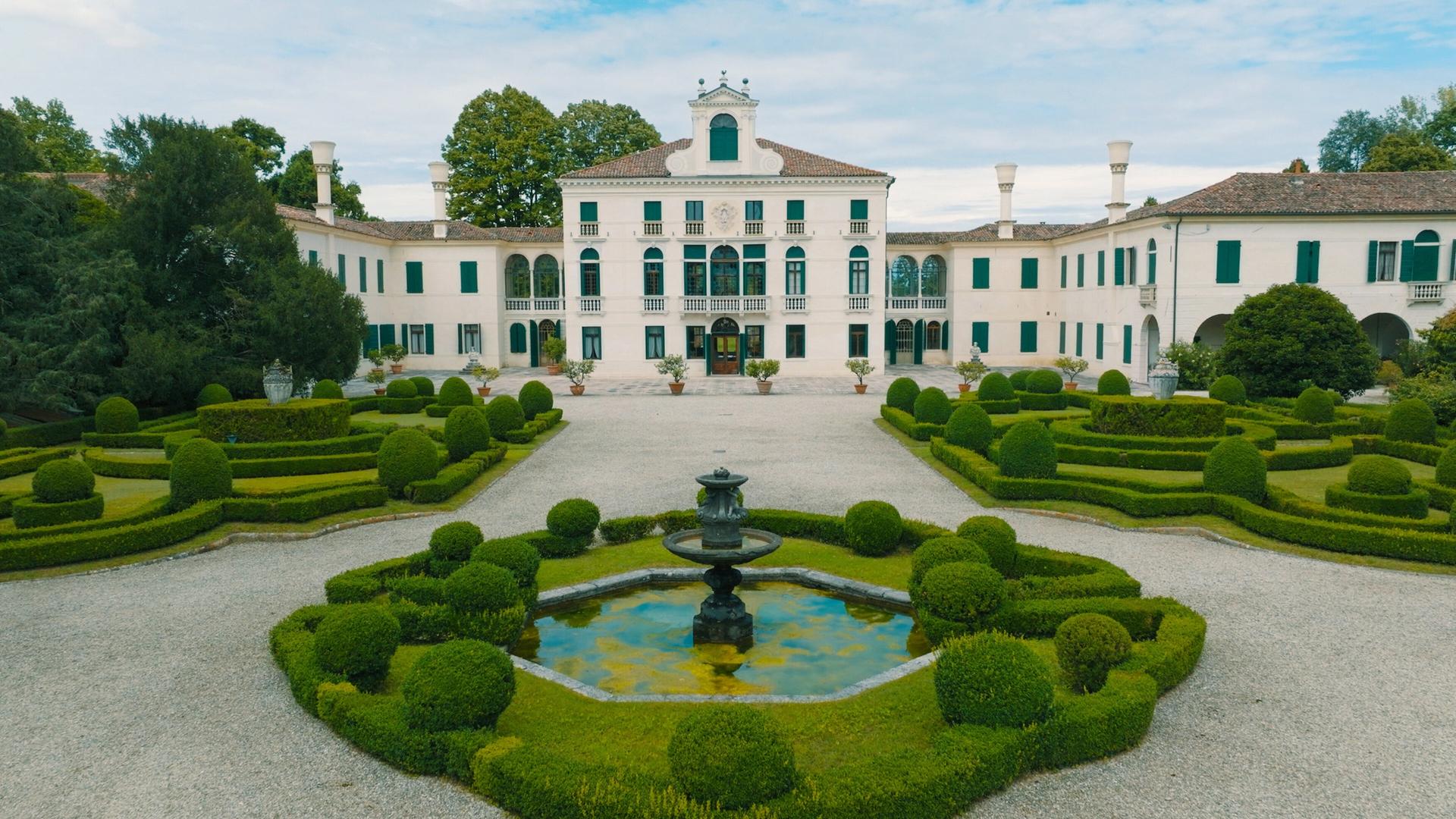 Das Bild zeigt die Villa Tiepolo Passi, eine prächtige Landvilla, umgeben von einem gepflegten Garten. Der zentrale Baukörper der Villa präsentiert sich in hellem Weiß mit eleganten Fenstern, die grüne Fensterläden besitzen. Auf dem Gebäude befindet sich ein zierlicher Dachaufsatz, der der Architektur einen klassischen Touch verleiht.  Vor der Villa erstreckt sich ein großzügiger Garten, der mit kunstvoll geschnittenen Hecken in runden und geschwungenen Formen gestaltet ist. In der Mitte des Gartens befindet sich ein kleiner Teich mit einer dekorativen Brunnenskulptur. Um den Teich sind ebenfalls grüne, üppige Pflanzen angeordnet, die einen harmonischen Gesamteindruck erzeugen.  Im Hintergrund sieht man Bäume, die den Garten umgeben und dem Bild eine natürliche Kulisse verleihen. Der Himmel ist leicht bewölkt, was für eine sanfte Beleuchtung sorgt, und die gesamte Szenerie strahlt eine ruhige, elegante Atmosphäre aus.