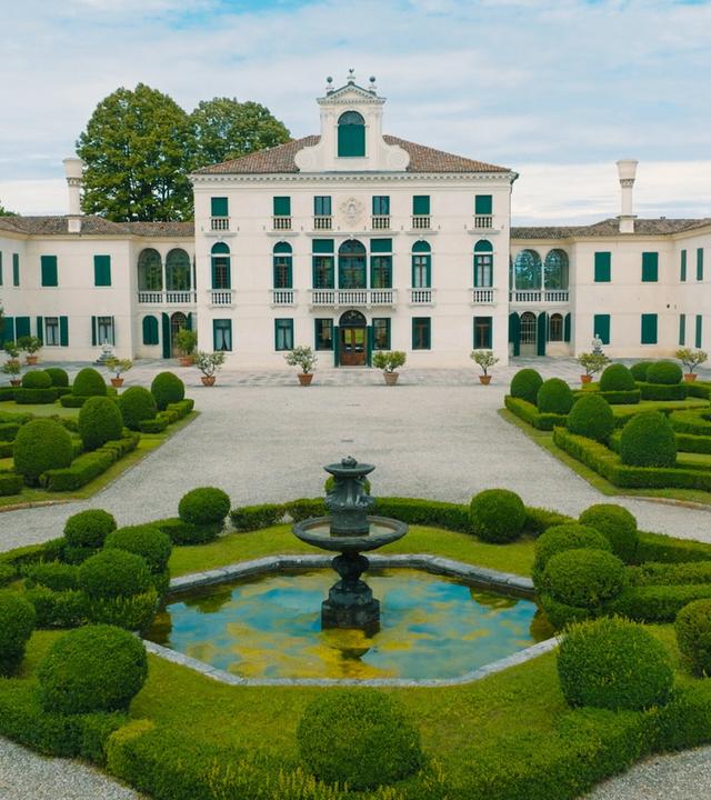Das Bild zeigt die Villa Tiepolo Passi, eine prächtige Landvilla, umgeben von einem gepflegten Garten. Der zentrale Baukörper der Villa präsentiert sich in hellem Weiß mit eleganten Fenstern, die grüne Fensterläden besitzen. Auf dem Gebäude befindet sich ein zierlicher Dachaufsatz, der der Architektur einen klassischen Touch verleiht.  Vor der Villa erstreckt sich ein großzügiger Garten, der mit kunstvoll geschnittenen Hecken in runden und geschwungenen Formen gestaltet ist. In der Mitte des Gartens befindet sich ein kleiner Teich mit einer dekorativen Brunnenskulptur. Um den Teich sind ebenfalls grüne, üppige Pflanzen angeordnet, die einen harmonischen Gesamteindruck erzeugen.  Im Hintergrund sieht man Bäume, die den Garten umgeben und dem Bild eine natürliche Kulisse verleihen. Der Himmel ist leicht bewölkt, was für eine sanfte Beleuchtung sorgt, und die gesamte Szenerie strahlt eine ruhige, elegante Atmosphäre aus.