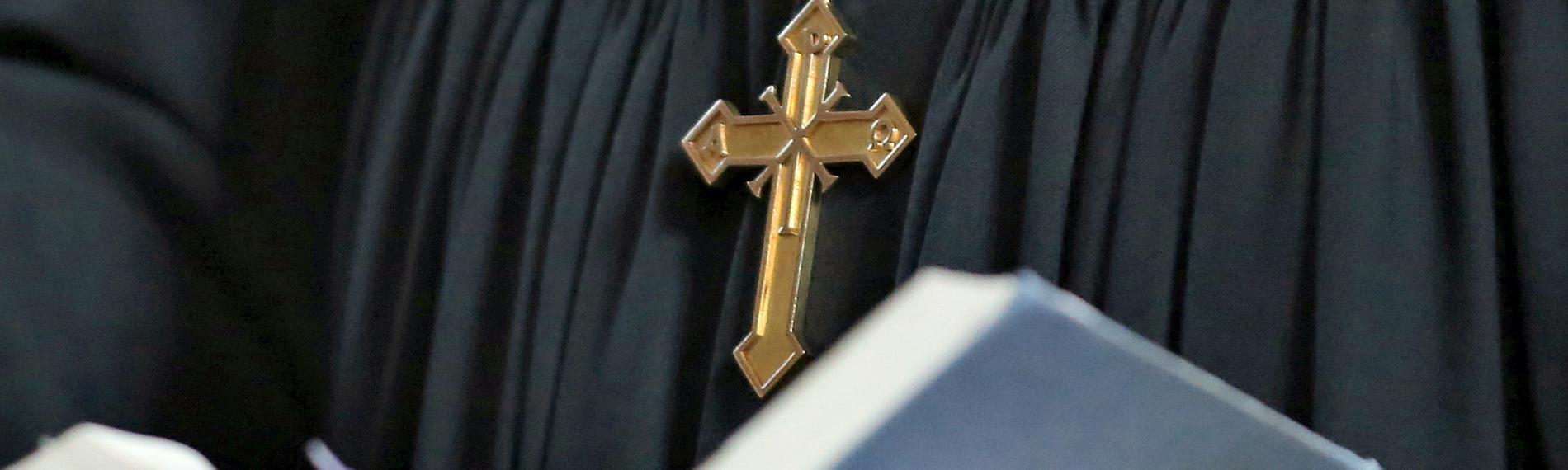 Ein Pfarrer mit Kreuzkette und schwarzem Gewand hält eine aufgeschlagene Bibel in seinen Händen.