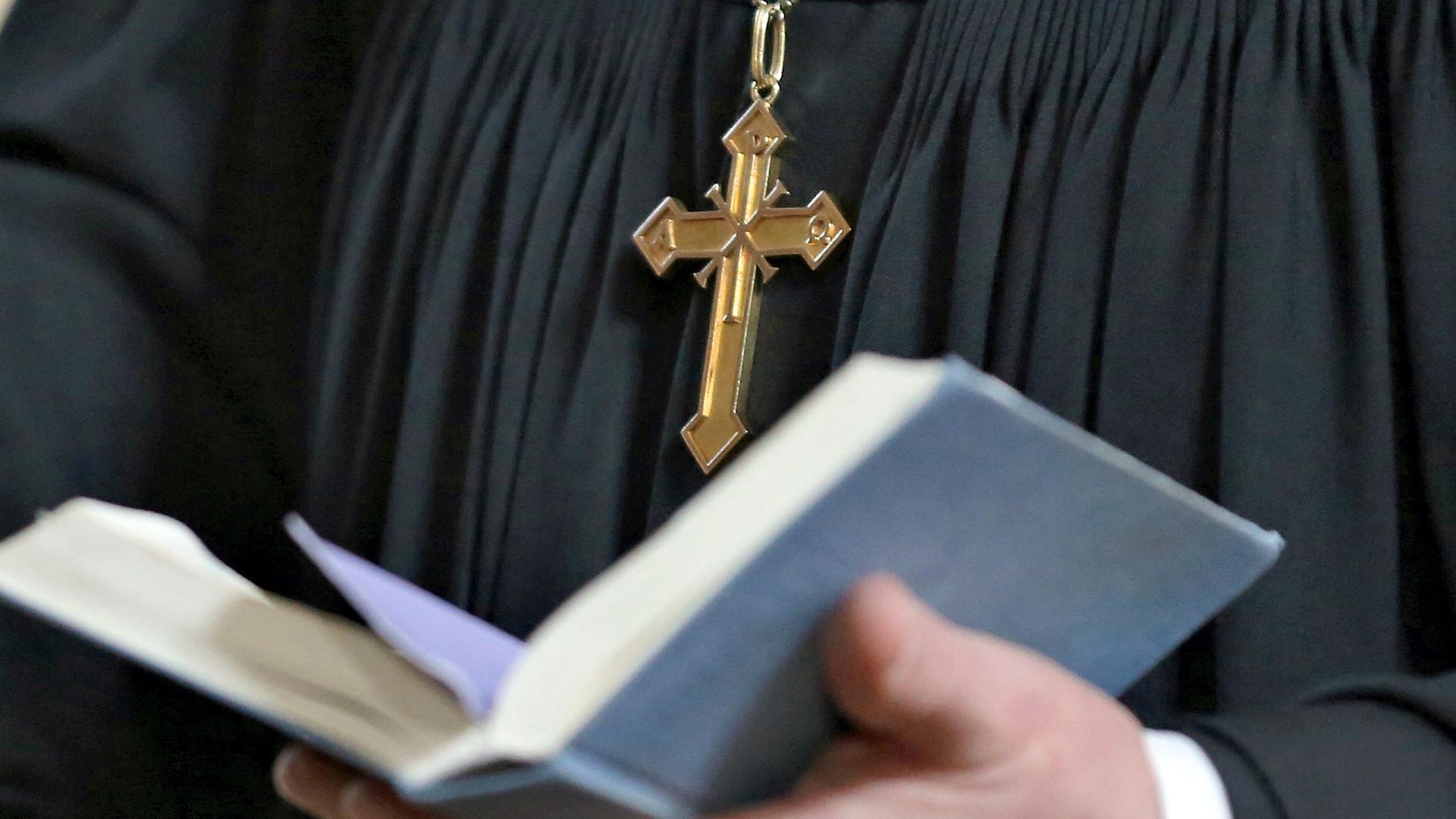 Ein Pfarrer mit Kreuzkette und schwarzem Gewand hält eine aufgeschlagene Bibel in seinen Händen.