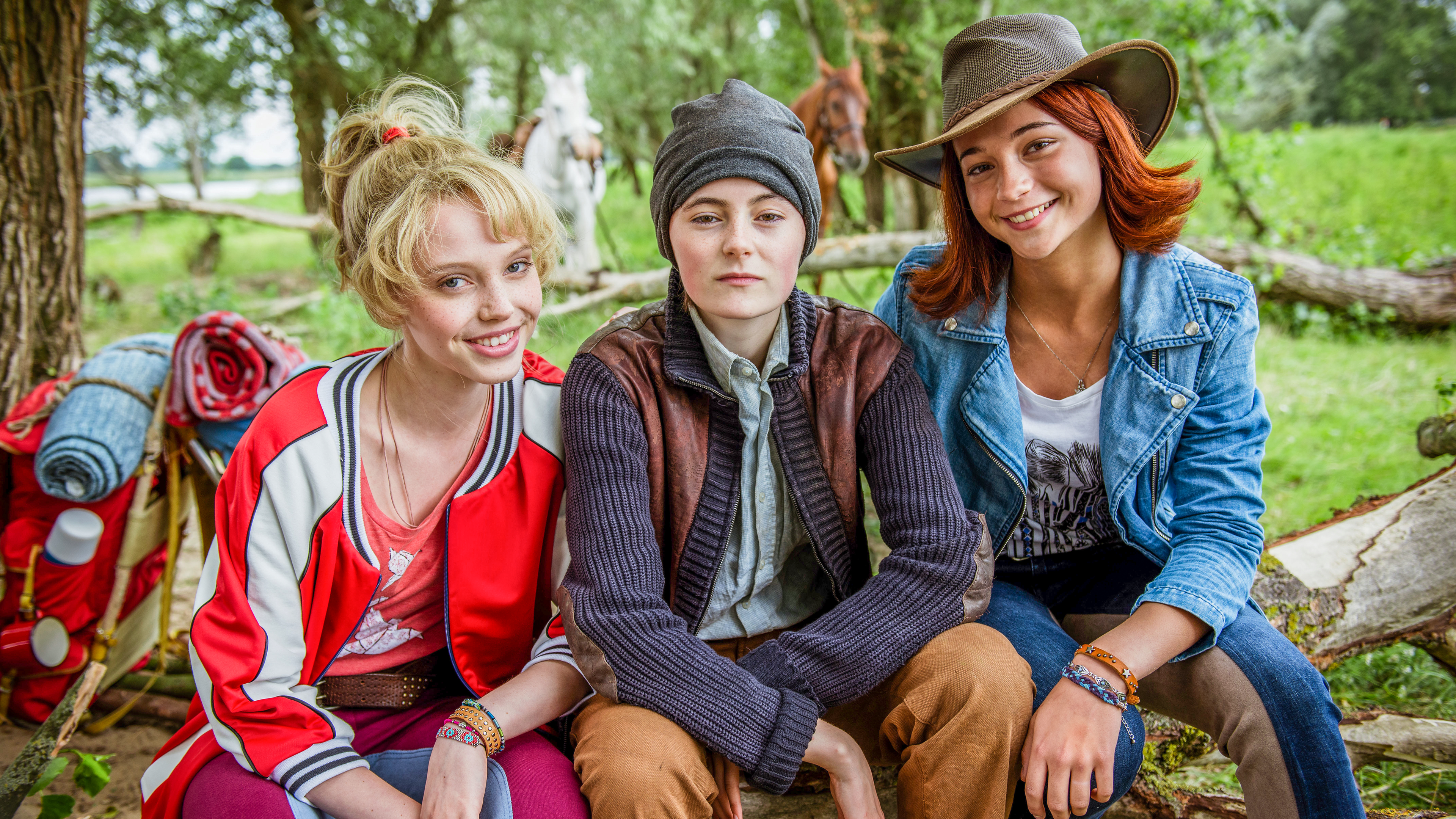 Die drei Mädchen Bibi (Lina Larissa Strahl), Adea/Aladin (Lea von Acken) und Tina (Lisa-Maire Koroll) sitzen nebeneinander auf einem Baumstamm. Bibi und Tina lächeln in die Kamera. Alea/Aladin trägt Männerkleidung und eine Mütze und blickt traurig in die Ferne. Im Hintergrund sind Bäume, zwei Pferde und eine grüne Wiese zu sehen.