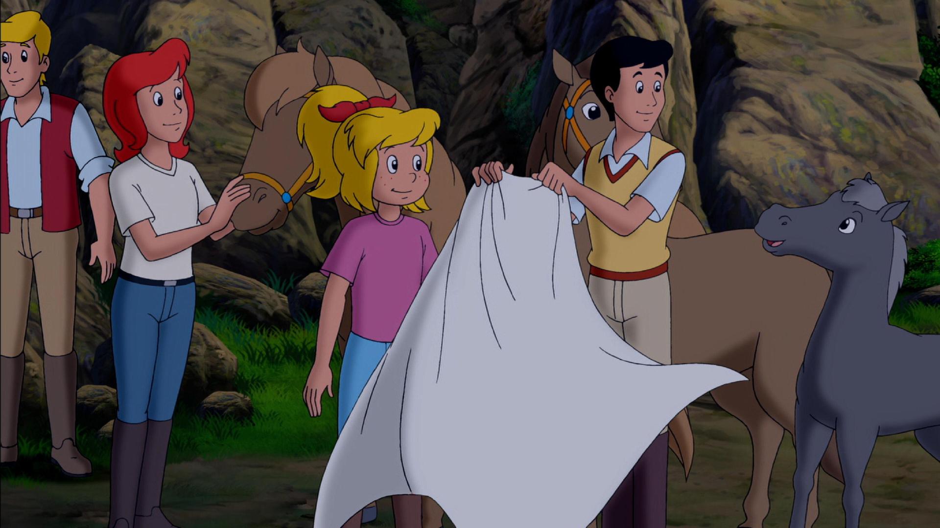 Bibi, Tina und Alex stehen vor einer felsigen Wand. Um sie herum tummeln sich drei Pferde, zwei größere und ein kleines. Alex hält in den Händen ein weißes Tuch, das im Wind weht.