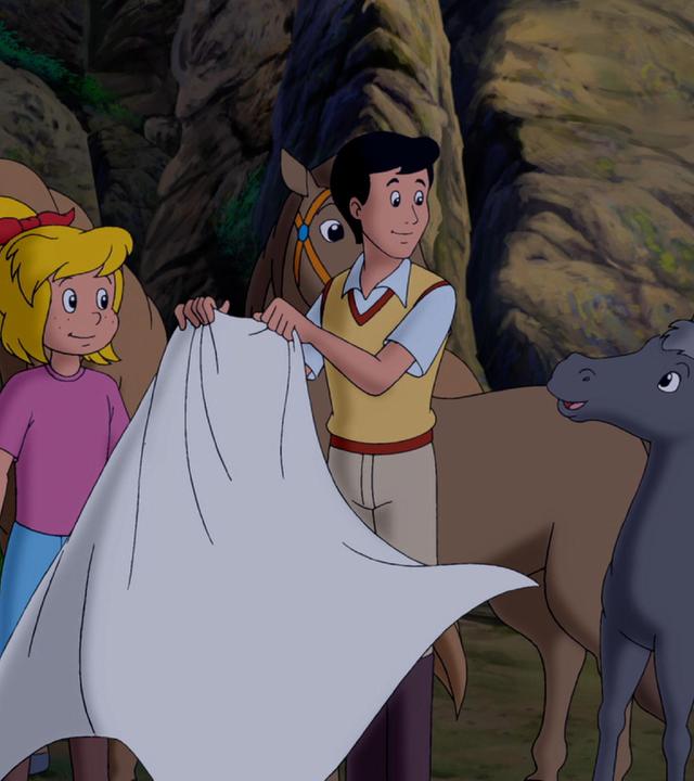 Bibi, Tina und Alex stehen vor einer felsigen Wand. Um sie herum tummeln sich drei Pferde, zwei größere und ein kleines. Alex hält in den Händen ein weißes Tuch, das im Wind weht.