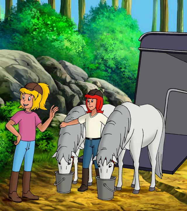 Bibi und Tina stehen mit den zwei Lipizzaner-Pferden vor dem Pferdetransporter. Die beiden Pferde fressen aus Eimern, die auf dem Boden stehen.