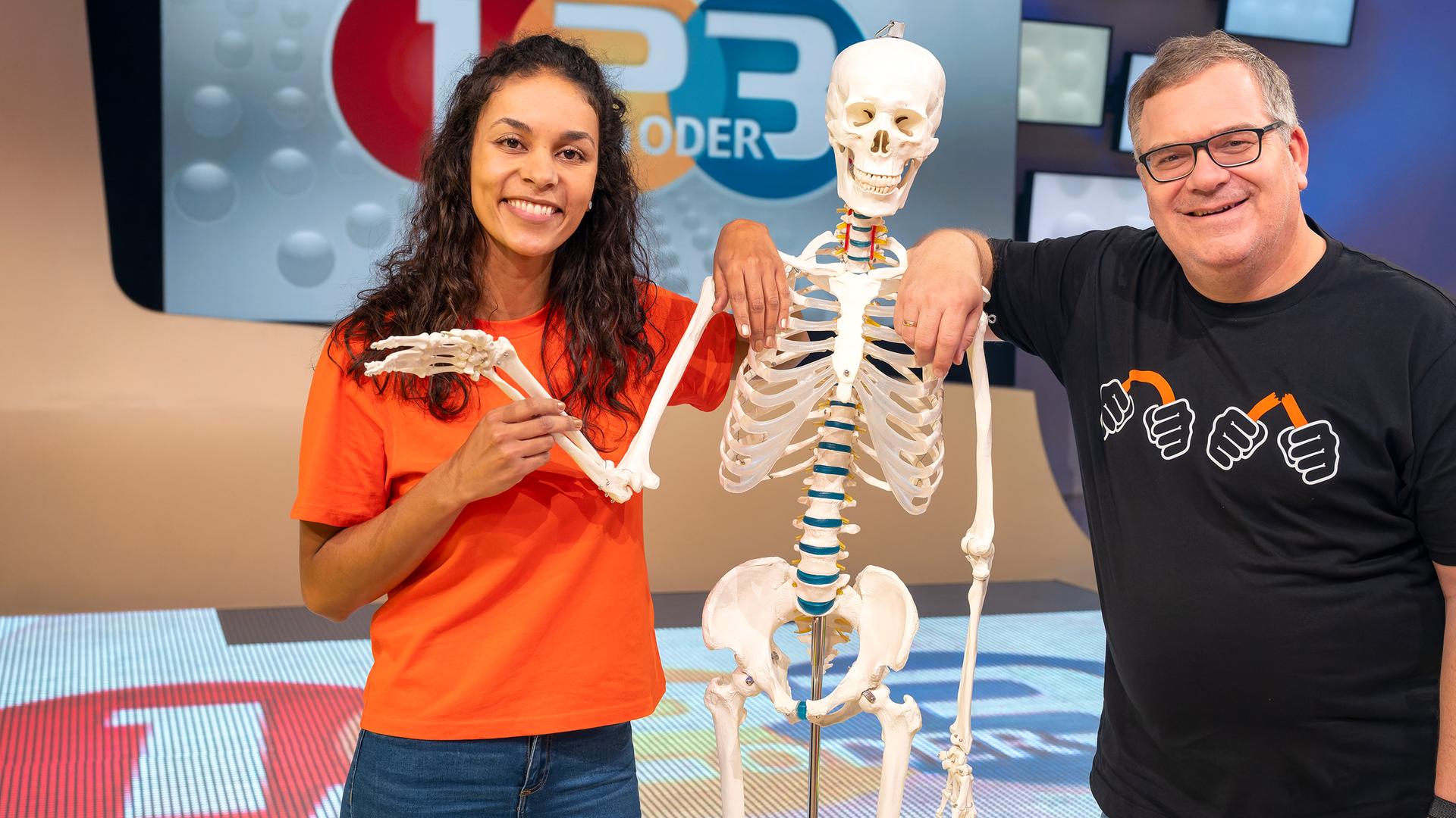 Studiogast Dr. Florence Randrianarisoa (l.) steht neben einem Skelett und hält den rechten Arm. Elton (l.) stützt sich auf die Schulter des Skeletts.