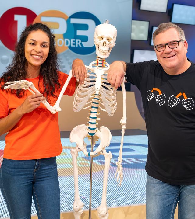 Studiogast Dr. Florence Randrianarisoa (l.) steht neben einem Skelett und hält den rechten Arm. Elton (l.) stützt sich auf die Schulter des Skeletts.