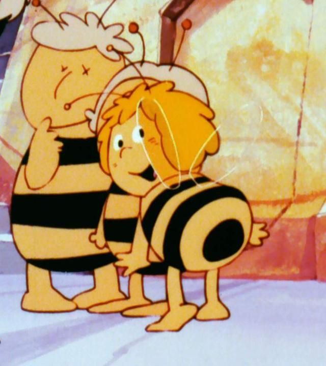 Die Biene Maja steht umringt von zwei weiteren kleinen Bienen und einer großen Biene im Mittelpunkt. Sie schaut fröhlich nach hinten zu ihren Flügeln. Die anderen schauen auf sie.
