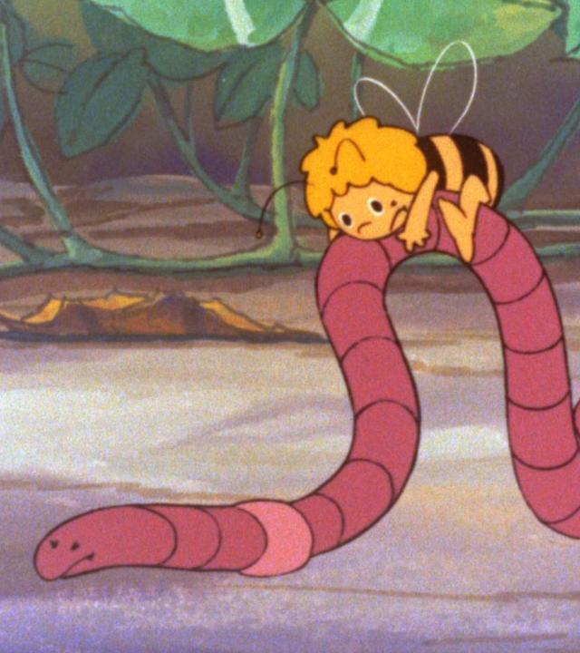 Bienenmädchen Maja sitzt auf einem Regenwurm, der sich in einer wellenartigen Bewegung fortbewegt. Maja hält sich krampfhaft fest.