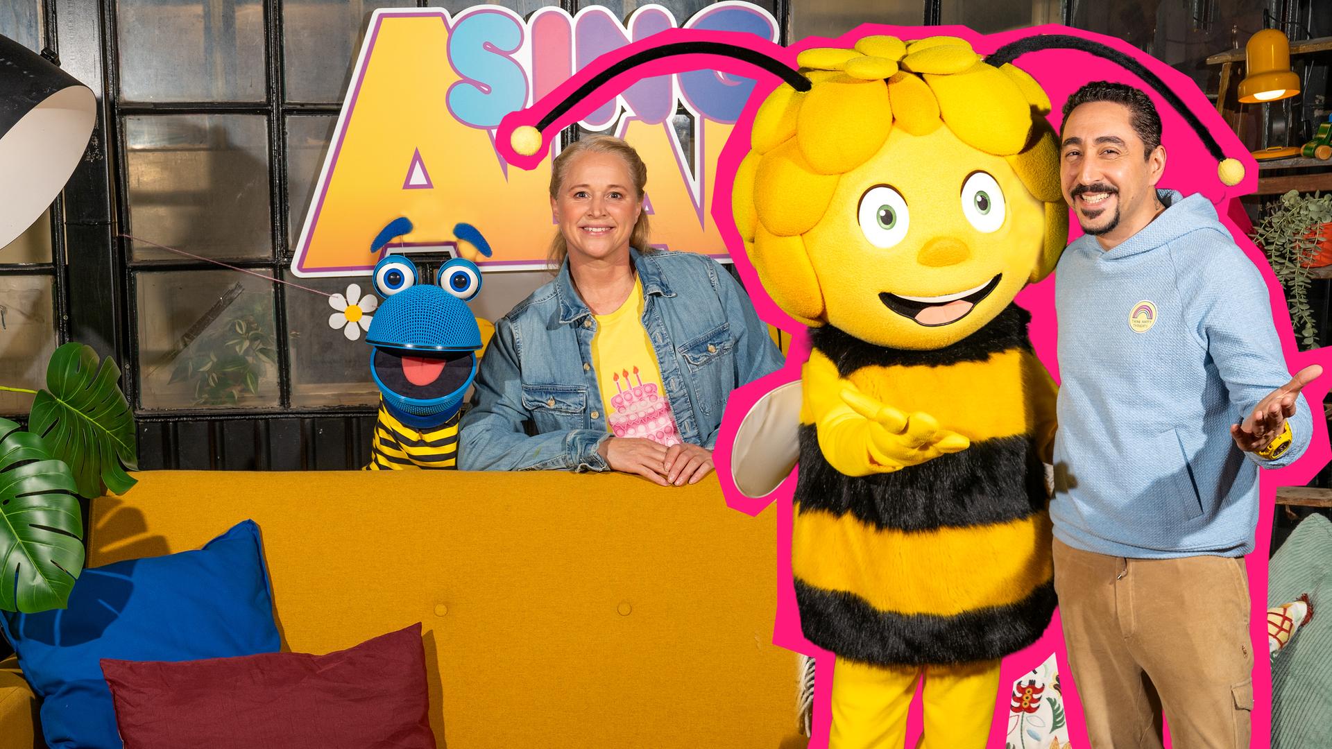 Das SingDing, Singa, Biene Maja und Eko Fresh sind auf dem Bild v. l. n. r. zu sehen - alle lächeln in die Kamera. SingDing trägt einen schwarz-gelb gestreiften Pullover befindet sich zusammen mit Singa hinter dem gelben Sofa. Eko und Maja stehen mit einladender Geste vor dem Sofa.