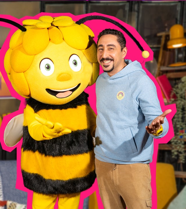 Das SingDing, Singa, Biene Maja und Eko Fresh sind auf dem Bild v. l. n. r. zu sehen - alle lächeln in die Kamera. SingDing trägt einen schwarz-gelb gestreiften Pullover befindet sich zusammen mit Singa hinter dem gelben Sofa. Eko und Maja stehen mit einladender Geste vor dem Sofa.