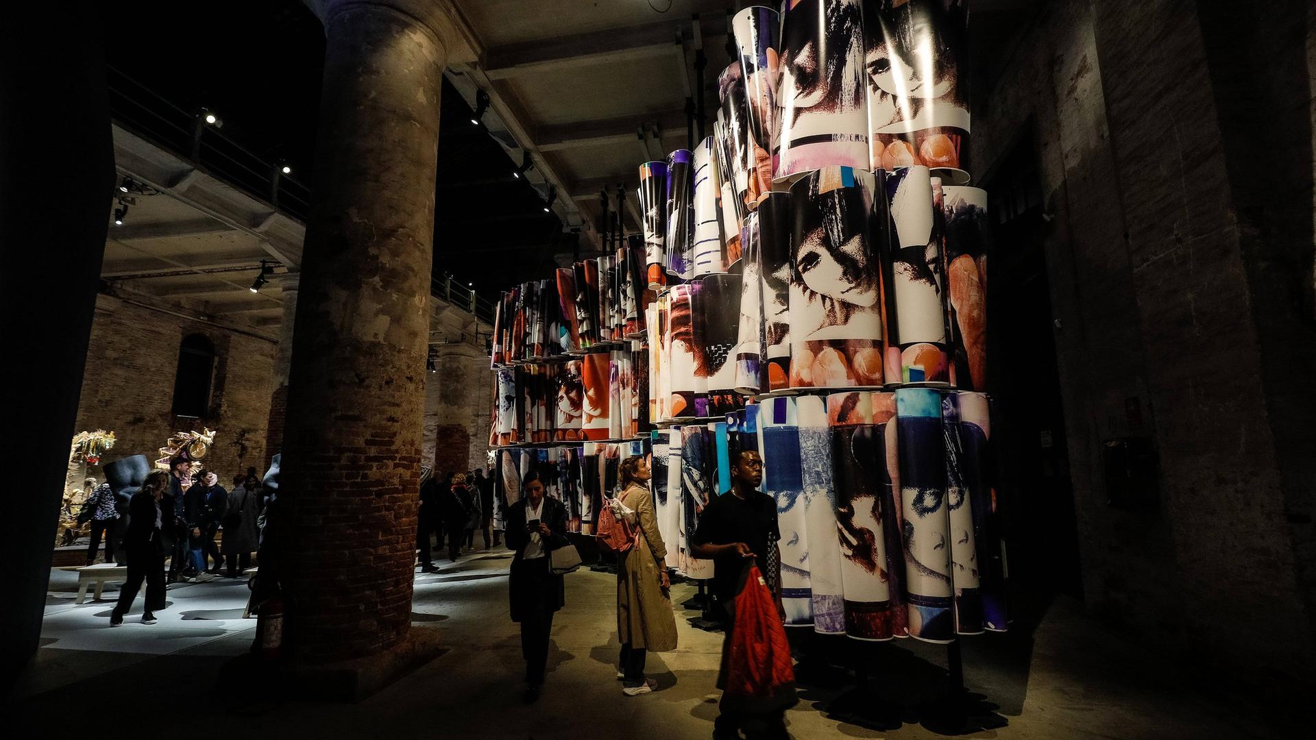 In einem halbdunklen Raum mit hoher Flachdecke laufen Menschen und betrachten ein riesiges Kunstwerk, das aus dreifach hoch gestapelten Bildrollen besteht.