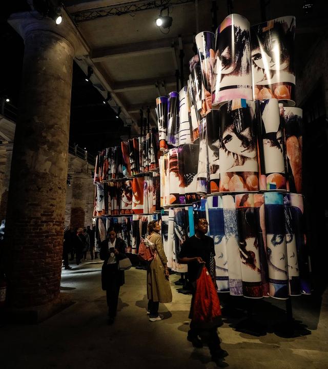 In einem halbdunklen Raum mit hoher Flachdecke laufen Menschen und betrachten ein riesiges Kunstwerk, das aus dreifach hoch gestapelten Bildrollen besteht.