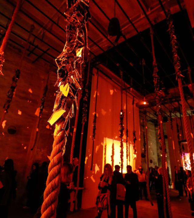 In einem halbdunklen Raum mit hoher Flachdecke laufen Menschen und betrachten ein riesiges Kunstwerk, das aus dreifach hoch gestapelten Bildrollen besteht.