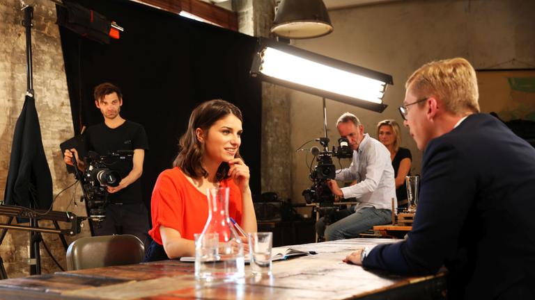  "Wähl mich! Jungpolitiker auf Stimmenfang": Sandra Rieß sitzt am Tisch im Gespräch mit einem Jungpolitiker, im Hintergrund ist ein Kamerateam zu sehen.