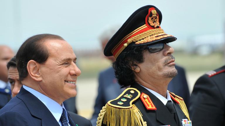 "Killing Gaddafi - Jagd auf den Diktator": Nahaufnahme von Gaddafi und Berlusconi. Sie stehen nebeneinander und schauen in eine Richtung.