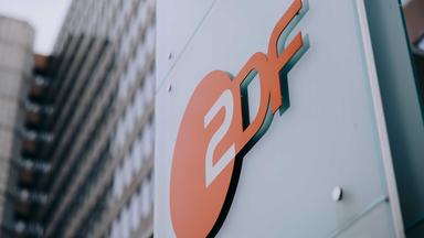 Nahaufnahme des ZDF-Logos vor moderner Hochhausfassade im Hintergrund. Das Bild repräsentiert das ZDF als Institution und wird als visuelles Element für Unternehmenskommunikation und Ausschreibungen eingesetzt.
