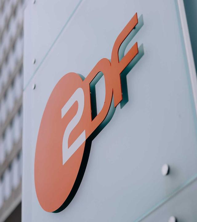 Nahaufnahme des ZDF-Logos vor moderner Hochhausfassade im Hintergrund. Das Bild repräsentiert das ZDF als Institution und wird als visuelles Element für Unternehmenskommunikation und Ausschreibungen eingesetzt.