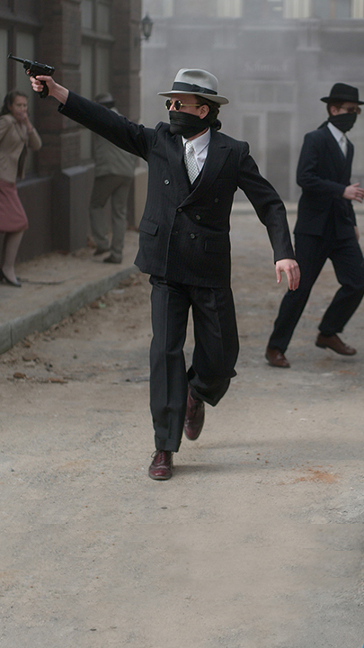 Presenter Sven Voss, im Hintergrund ein nachgestelltes Foto eines Überfalls. Mehrere Männer mit Maske und Sonnenbrille, ein Mann hält eine Pistole in der Hand.