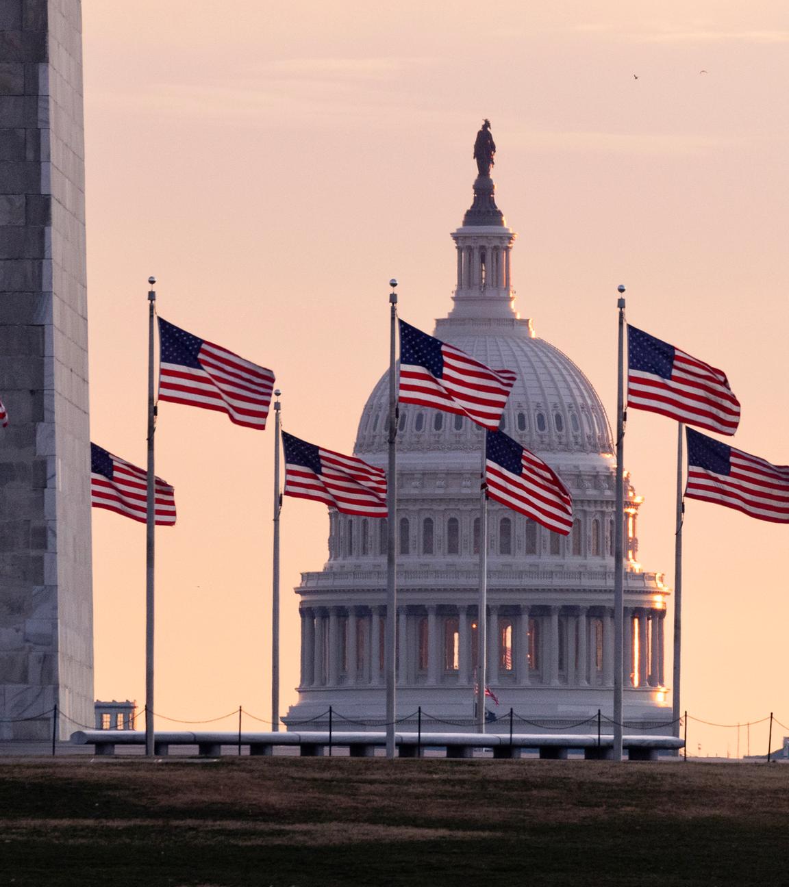 Im Hintergrund geht über dem Kapitol der Vereinigten Staaten von Amerika die Sonne auf, im Vordergrund sind US-Flaggen am Washington Monument zu sehen. 