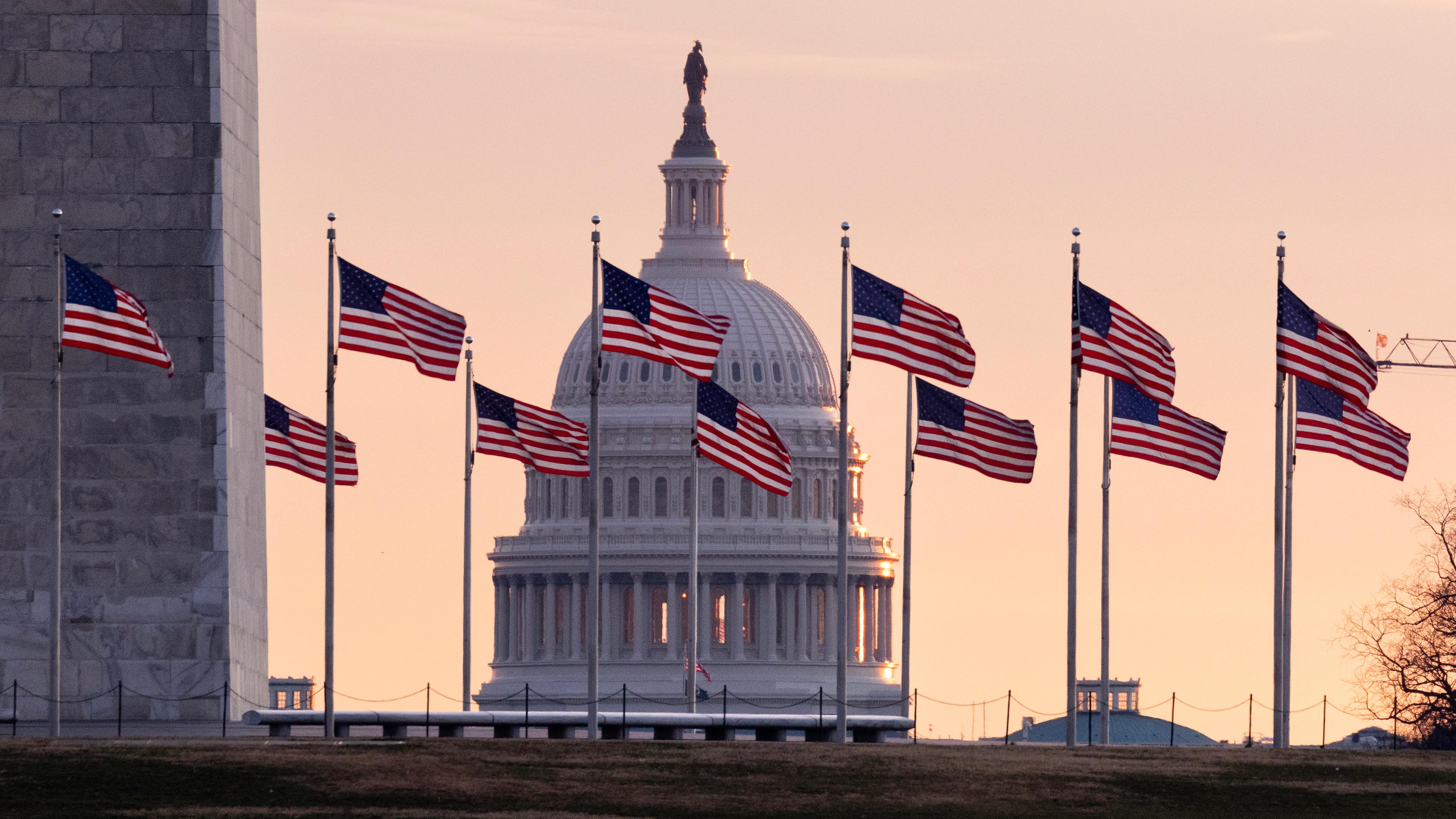 Im Hintergrund geht über dem Kapitol der Vereinigten Staaten von Amerika die Sonne auf, im Vordergrund sind US-Flaggen am Washington Monument zu sehen. 