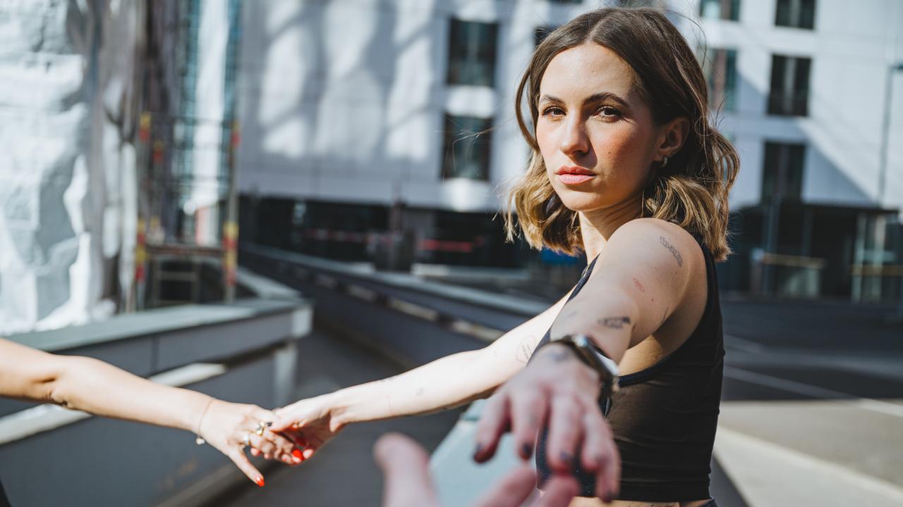 Eine junge Frau entfernt sich mit ausgestrecktem Arm von einer Person außerhalb des Bildrandes und streckt den anderen Arm nach einer dritten Person außerhalb des Bildrandes aus.