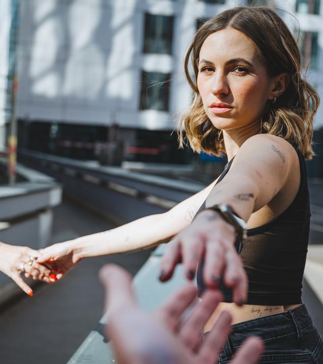 Eine junge Frau entfernt sich mit ausgestrecktem Arm von einer Person außerhalb des Bildrandes und streckt den anderen Arm nach einer dritten Person außerhalb des Bildrandes aus.