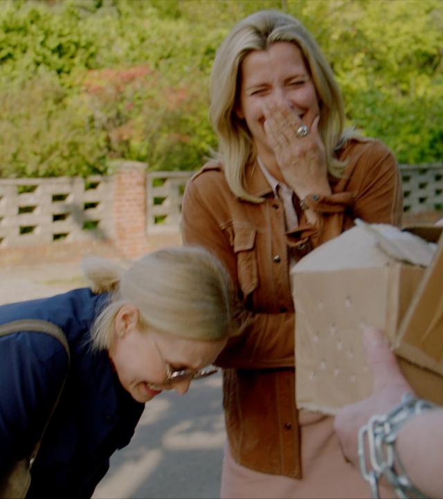 Drei Personen im Freien: Zwei lachen herzlich, eine hält sich die Hand vor den Mund, die andere beugt sich vor. Im Vordergrund steht eine dritte Person mit einem Paket in den Händen. Im Hintergrund sind grüne Pflanzen und eine gemauerte Wand zu sehen