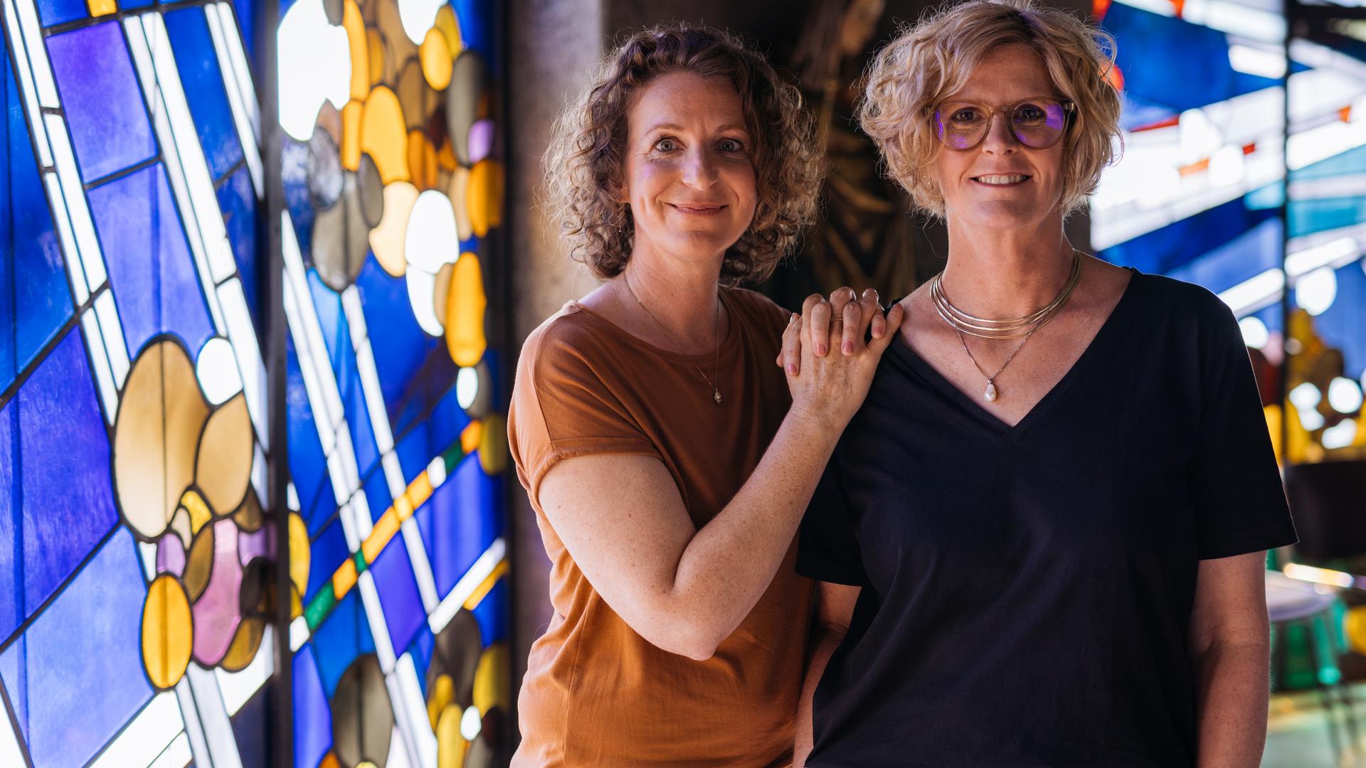 Nele und Kerstin stehen nebeneinander, die Hände von Nele liegen auf Kerstins Schulter. Beide lächeln freundlich in die Kamera. Im Hintergrund sind bunte Kirchenfenster zu sehen.