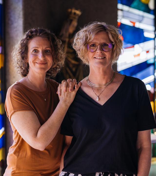 Nele und Kerstin stehen nebeneinander, die Hände von Nele liegen auf Kerstins Schulter. Beide lächeln freundlich in die Kamera. Im Hintergrund sind bunte Kirchenfenster zu sehen.