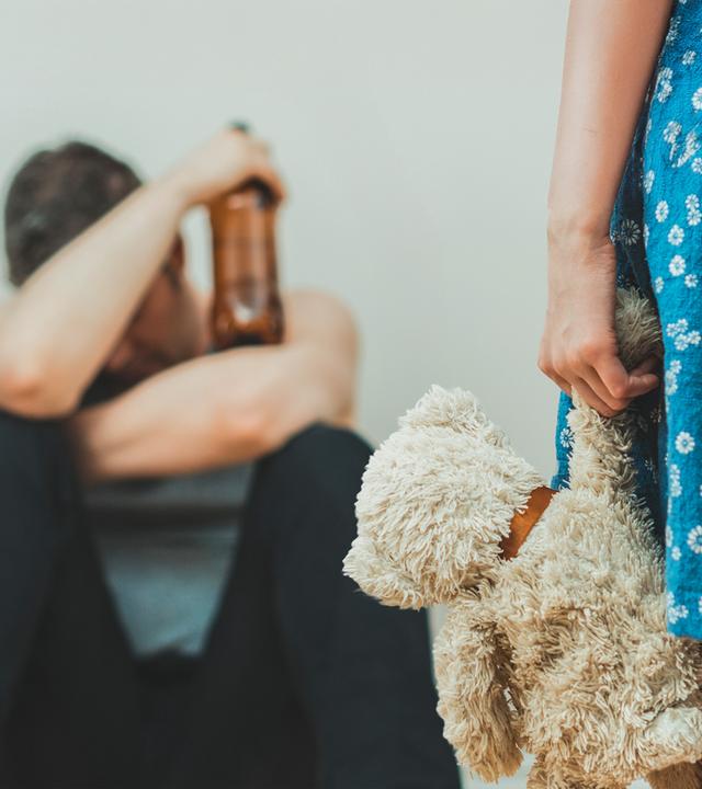 Vater sitzt mit Bier auf dem Boden und Mädchen mit Teddy schaut zu