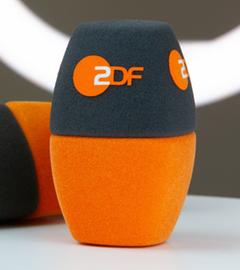 Im Vordergrund sind zwei zweifarbige ZDF-Mikrofon-Windschutzaufsätze zu sehen, einer aufrecht, einer liegend. Die Aufnahme symbolisiert journalistische Arbeit und den Pressebereich des ZDF.