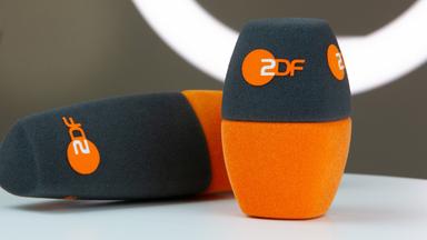 Im Vordergrund sind zwei zweifarbige ZDF-Mikrofon-Windschutzaufsätze zu sehen, einer aufrecht, einer liegend. Die Aufnahme symbolisiert journalistische Arbeit und den Pressebereich des ZDF.