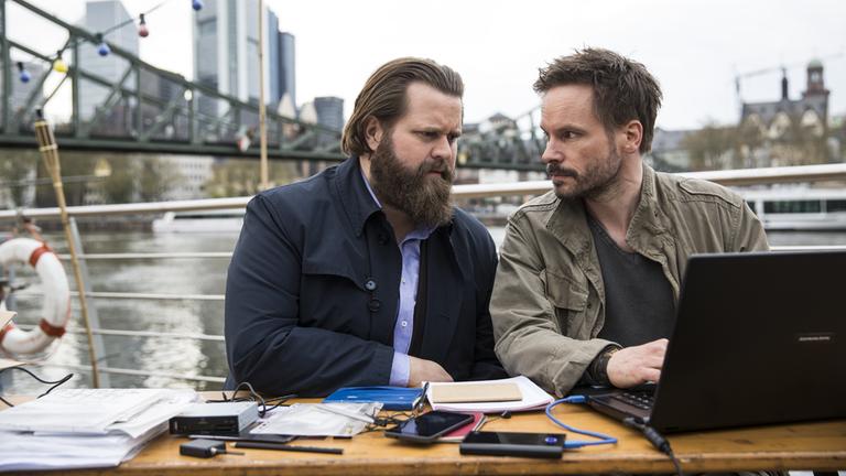 "Ein Fall für zwei - Das schwarze Schaf": Benni Hornberg (Antoine Monot, Jr.) und Leo Oswald (Wanja Mues) sitzen an Deck von Leos Hausboot. Leo hat einen Laptop vor sich und schaut Benni an. Im Hintergrund die Frankfurter Skyline.