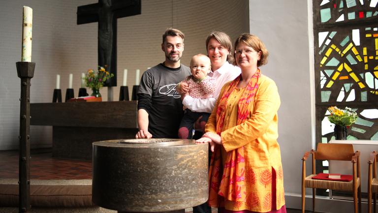 "37°: Mutter, Mutter, Vater, Kind - Elternglück für Lesben und Schwule": Vater Nils steht mit den beiden Müttern vor dem Taufbecken in der Kirche. Mutter Annika hält das gemeinsame Kind im Arm.