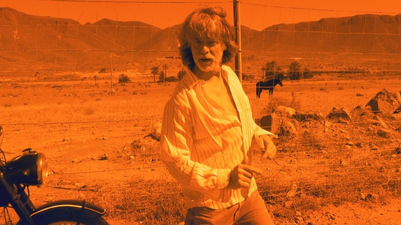 "Mülheim – Texas. Helge Schneider hier und dort": Auf einem stark orange eingefärbten Bild tanzt Helge Schneider, vor einer Wüstenlandschaft. Neben ihm ist ein Motorrad zu sehen.