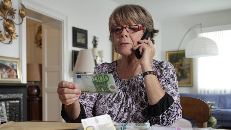 "Aktenzeichen XY-Spezial: Vorsicht, Betrug!": Eine Frau sitzt am Schreibtisch und hält einen Hundert Euro Schein in der Hand. Vor ihr liegen noch mehrere Bündel Geldscheine.