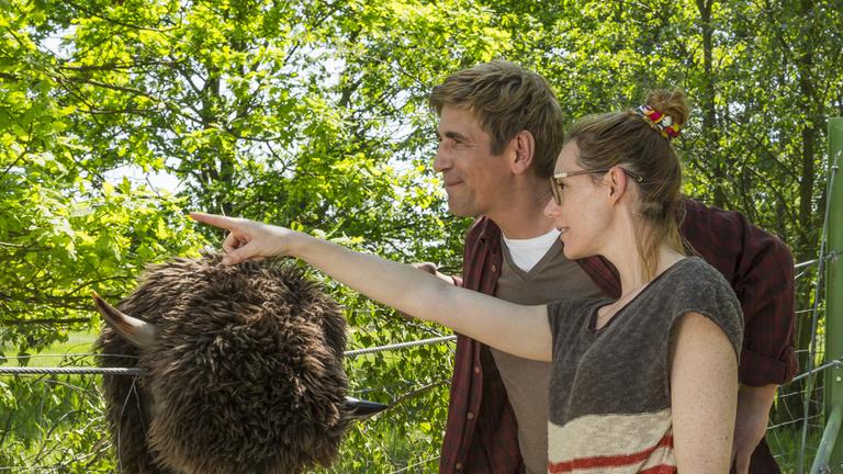 "Löwenzahn - Wisent - Urtiere in Gefahr": Fritz Fuchs (Guido Hammesfahr) steht mit der Leiterin des Naturparks (Lavinia Wilson) vor einem Zaun, auf dem ein braunes Wisent-Kostüm hängt. Die Leiterin zeigt mit dem Finger in die Ferne.