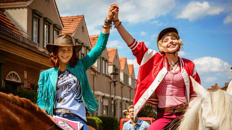 "Bibi & Tina - Voll verhext": Im Hintergrund eine Dorfstraße. Im Vordergrund Tina (Lisa-Marie Koroll) und Bibi (Lina Larissa Strahl). Beide sitzen auf ihren Pferden, haben sich an der Hand gepackt und strecken die beiden Arme steil in die Höhe. Siegessicher lächeln sie in die Kamera.
